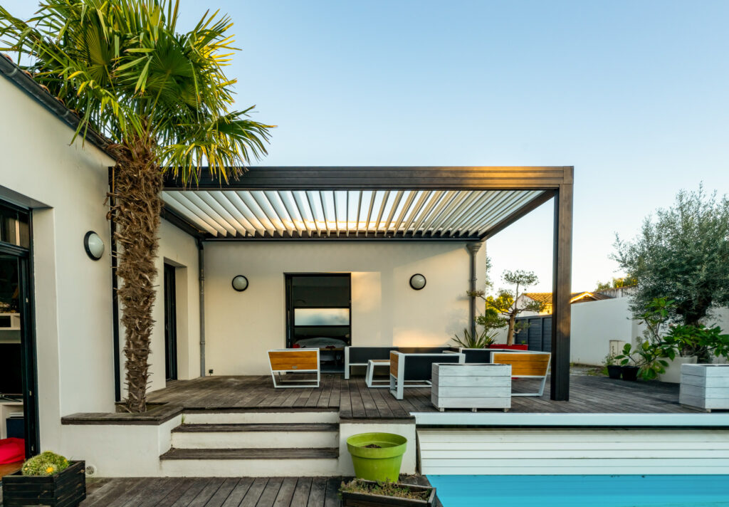 : Pergola noire aux lames fermées installée sur une terrasse en bois avec un palmier.